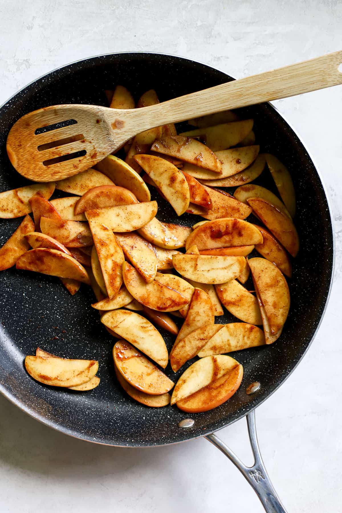 Simple sautéed apples in maple syrup and cinnamon, in black sauté pan. A bamboo spoon is resting on the side of the pan.