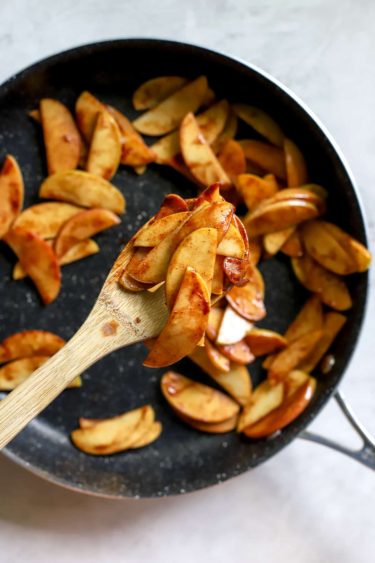 Wooden spoon full of simple sautéed apples, above sauté pan with more apples.