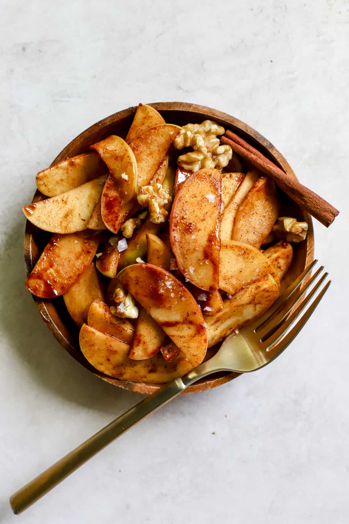 Simple sautéed apples on small wooden plate, topped with walnuts and flaky salt, with a golden fork on gray/white surface.