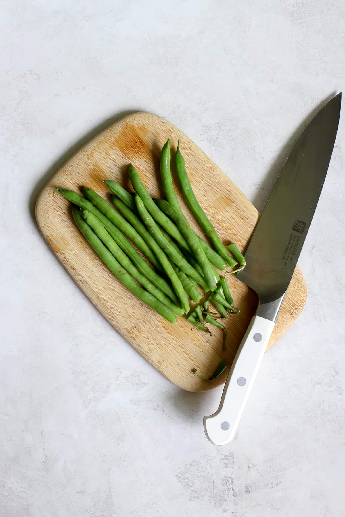 Green beans being trimmed on small wooden cutting board with chef's knife, on gray and white surface.