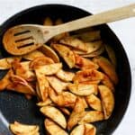 Sautéed apples with maple syrup and cinnamon in dark gray pan with wooden spoon.