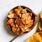 Simple sautéed apples on small wooden plate, topped with walnuts and flaky salt, with a golden fork on gray/white surface with golden yellow linen