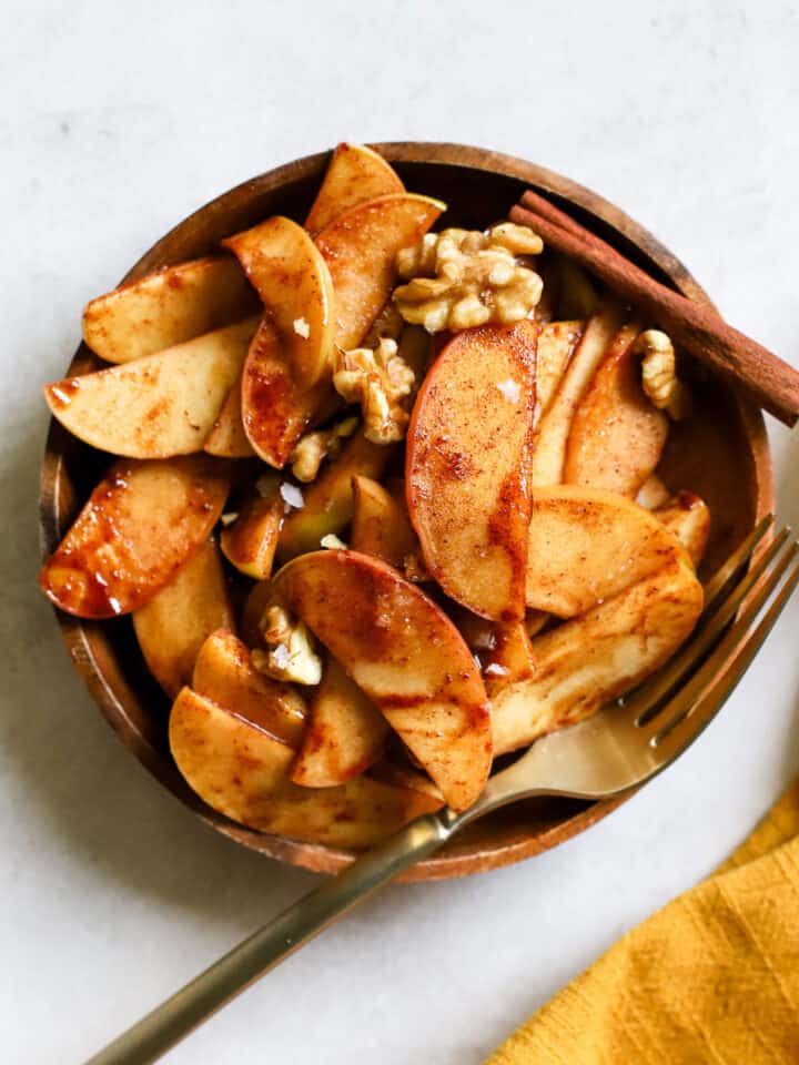 Simple sautéed apples on small wooden plate, topped with walnuts and flaky salt, with a golden fork on gray/white surface with golden yellow linen