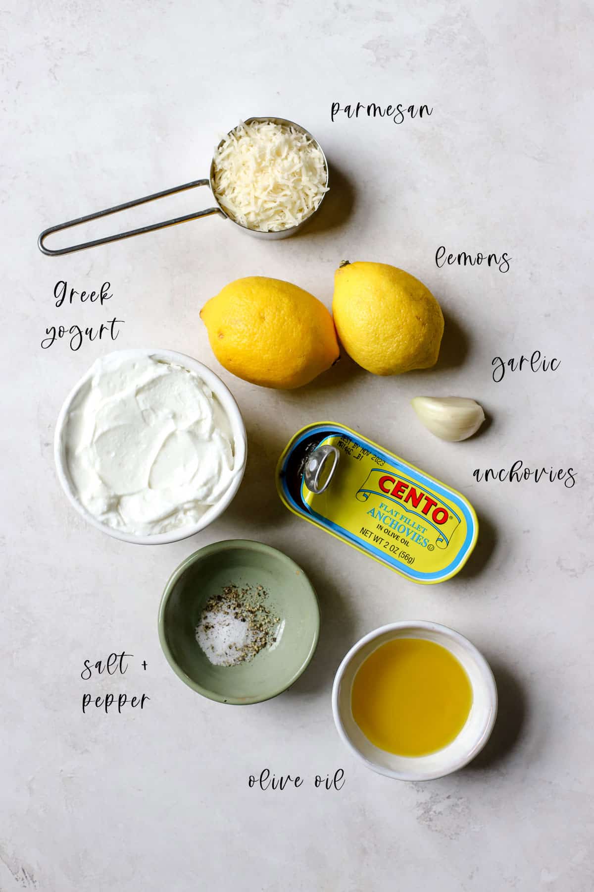 Grated parmesan in a measuring cup, two lemons, a garlic clove, a small white bowl with Greek yogurt, a can of Cento anchovies packed in olive oil, a small green bowl of salt and pepper, and a small white bowl with olive oil are the ingredients needed to Caesar dressing, sitting in a gray and white surface.