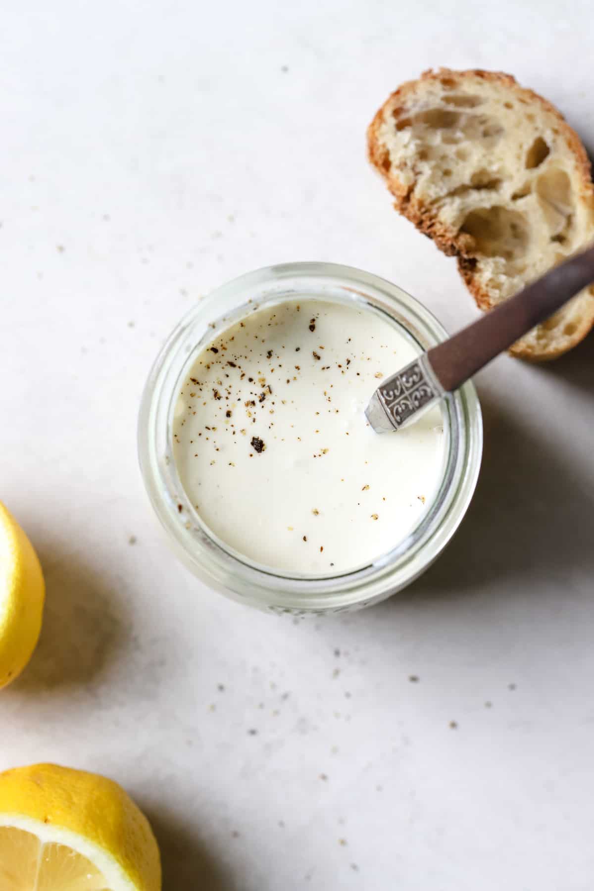Homemade Caesar dressing is in a small jar with freshly cracked pepper on top and spoon resting in the jar on a light gray surface. Next to the dressing are a couple lemon slices and a slice of fresh sourdough bread.