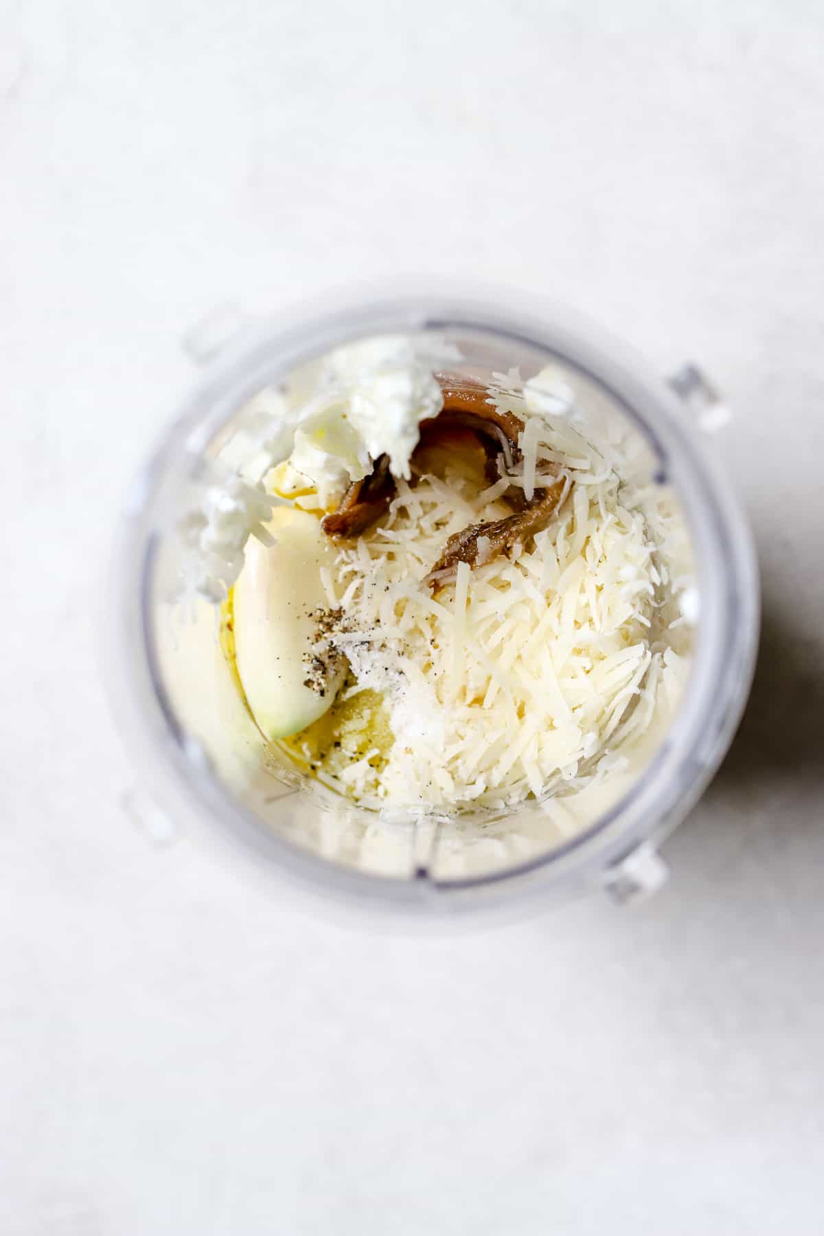 Homemade Caesar dressing ingredients in a small blender cup before being blended, on a gray and white surface.