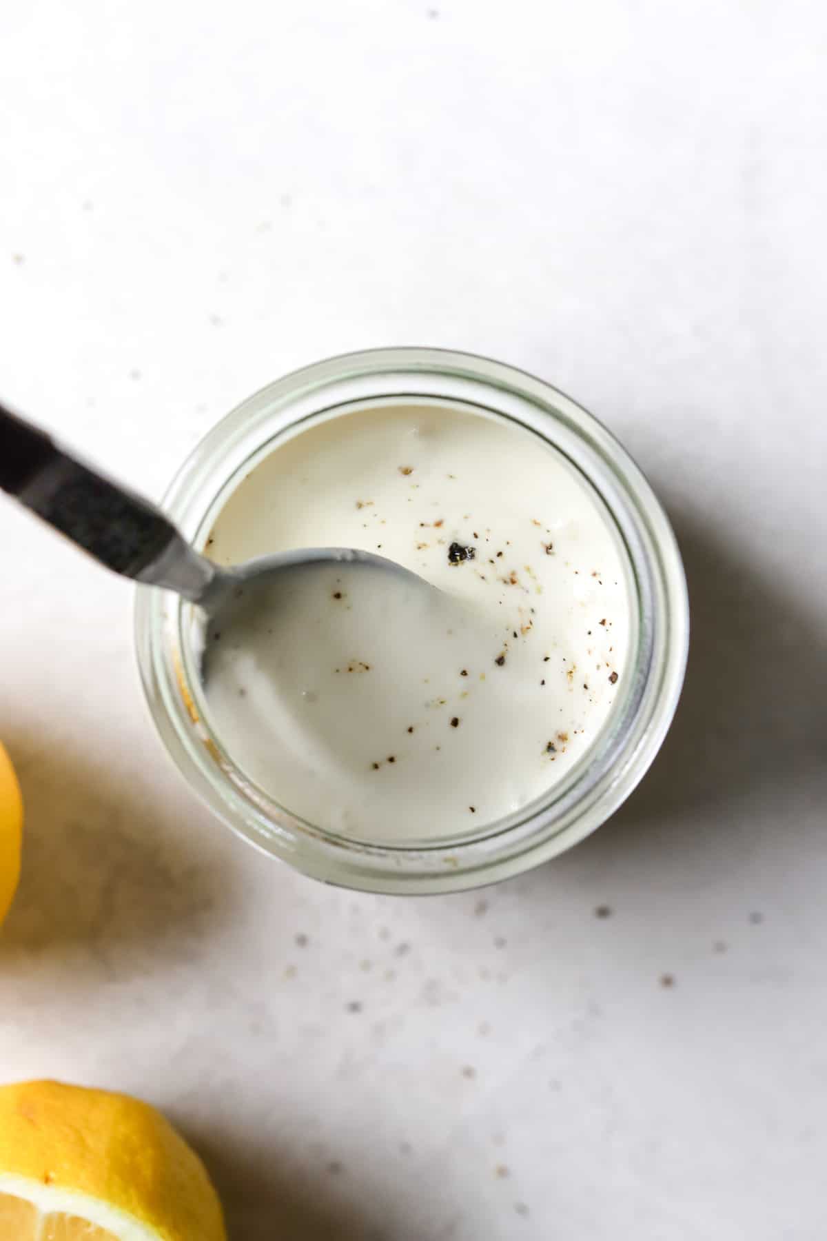 Caesar dressing in small glass jar with freshly cracked pepper on top, being lightly stirred by a spoon, on light gray surface with lemons on side