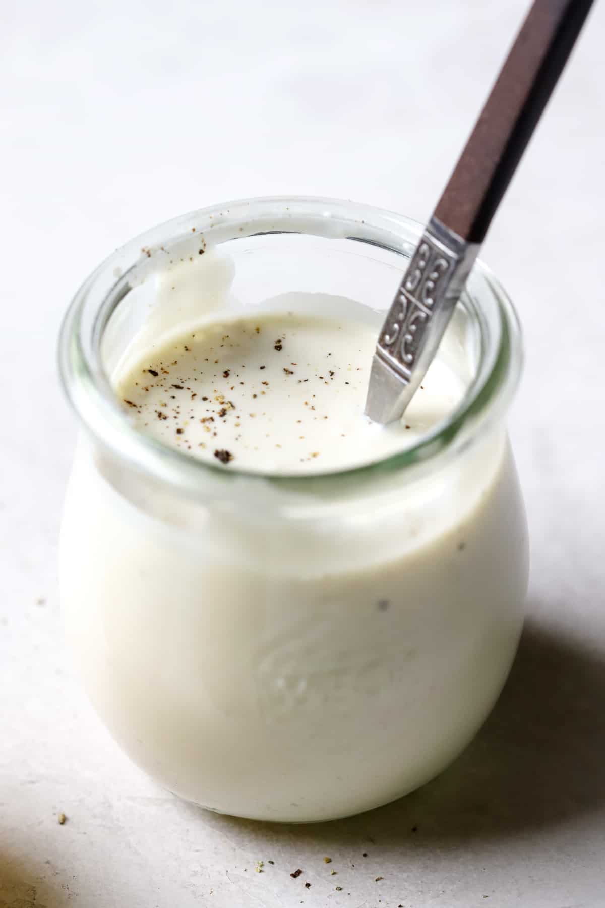 Caesar dressing in small glass jar being lightly stirred by a spoon, on light gray surface.