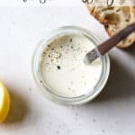 Homemade Caesar dressing in a small jar with freshly cracked pepper on top, on light gray surface, with slice of sourdough and lemons on side