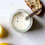 Homemade Caesar dressing in a small jar with freshly cracked pepper on top, on light gray surface, with slice of sourdough and lemons on side