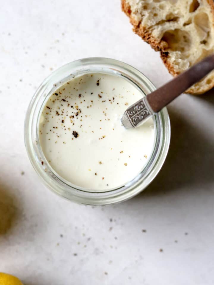 Homemade Caesar dressing in a small jar with freshly cracked pepper on top, on light gray surface, with slice of sourdough and lemons on side