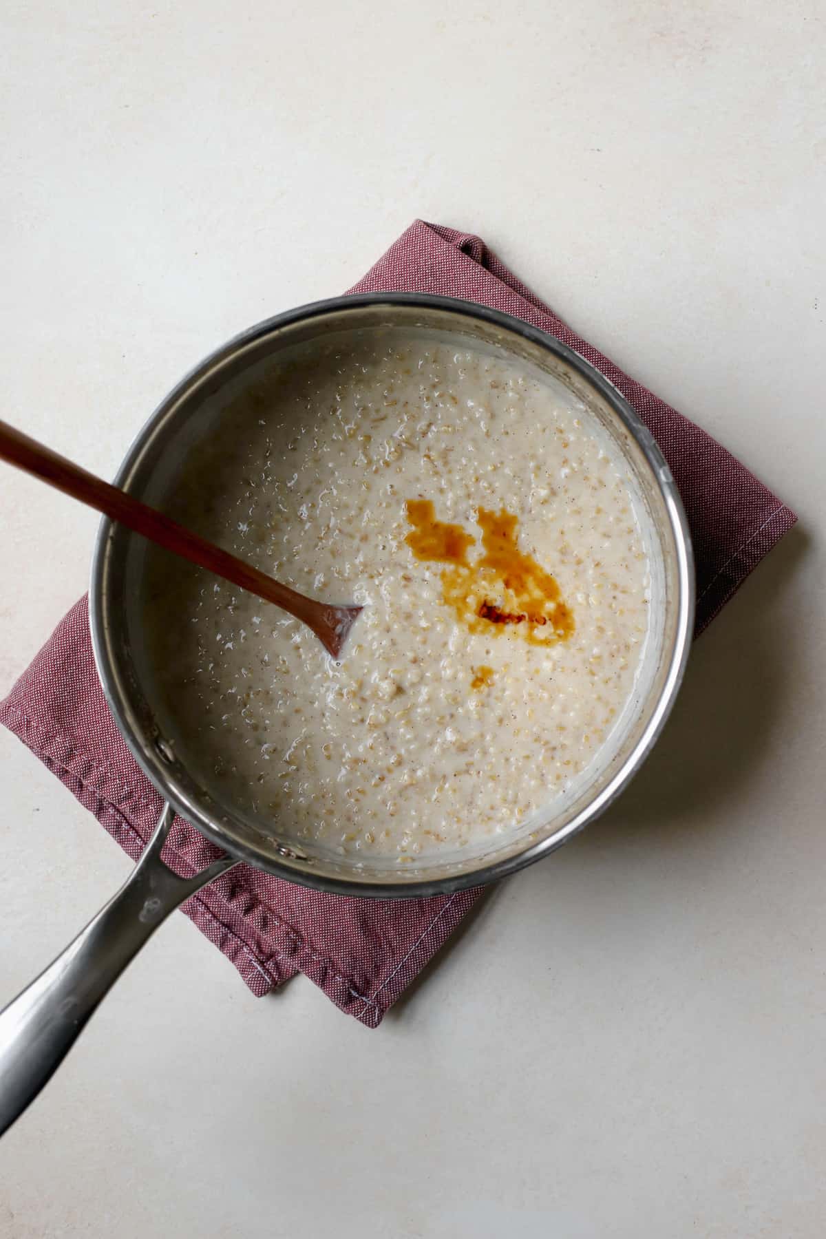 Fully cooked steel cut oats in stainless steel saucepan with a splash of vanilla added, with wooden spoon.