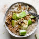 Steel cut oats in a small bowl with green apple chunks, walnuts, cinnamon, and maple syrup drizzled over the top