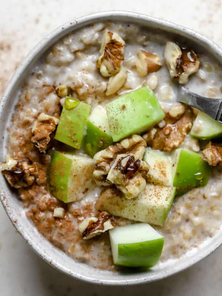 Steel cut oats in a small bowl with green apple chunks, walnuts, cinnamon, and maple syrup drizzled over the top