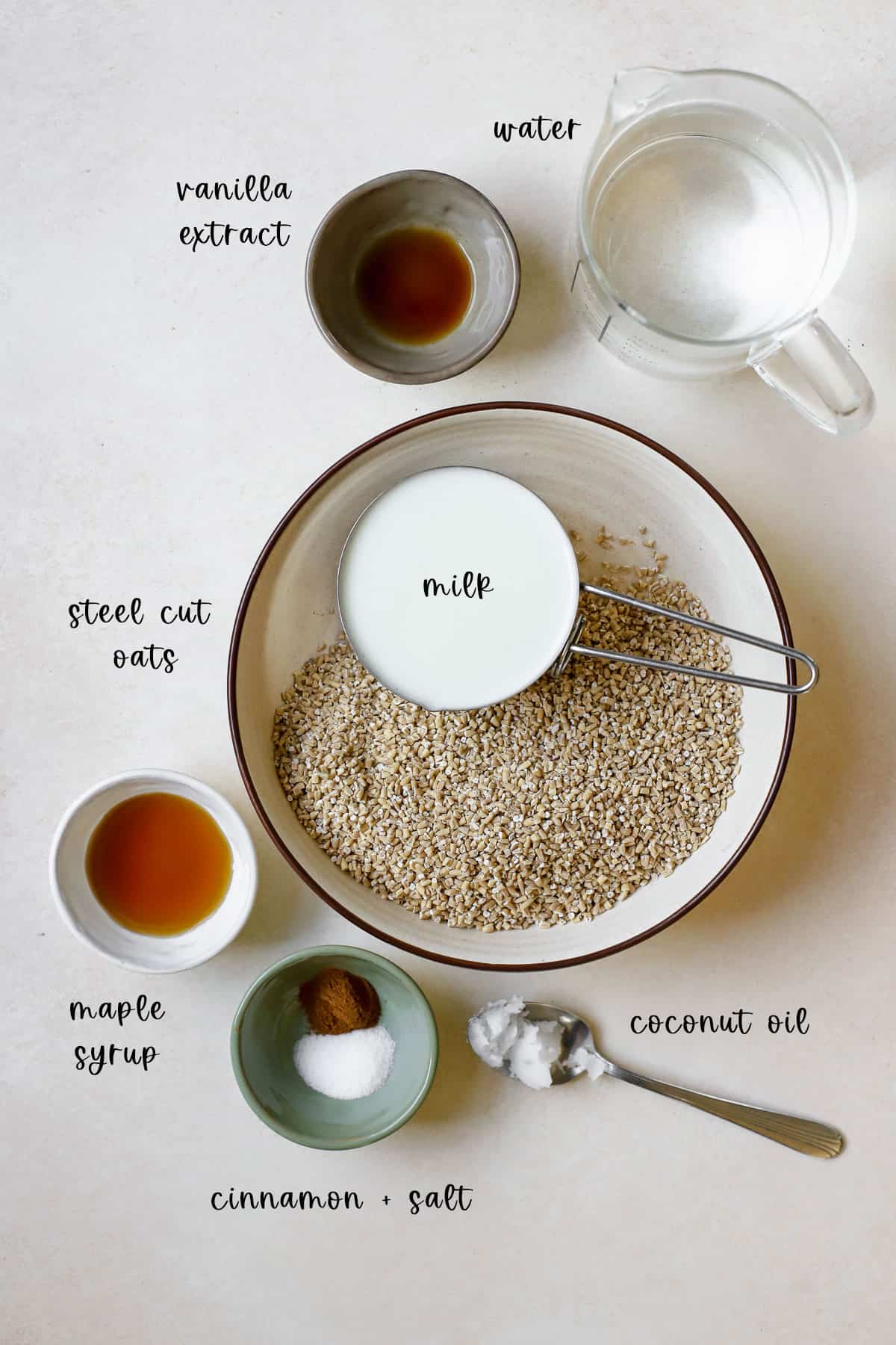 Steel cut oats ingredients including steel cut oats, milk, water, coconut oil, maple syrup, vanilla extract, cinnamon, and salt, all measured into various sized bowls on beige surface.