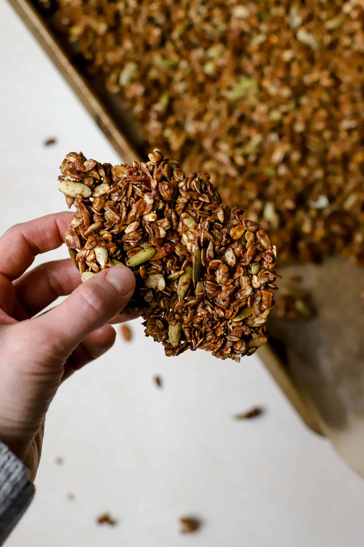 Hand holding a large flat chunk of ginger molasses granola, with sheet pan of granola in background