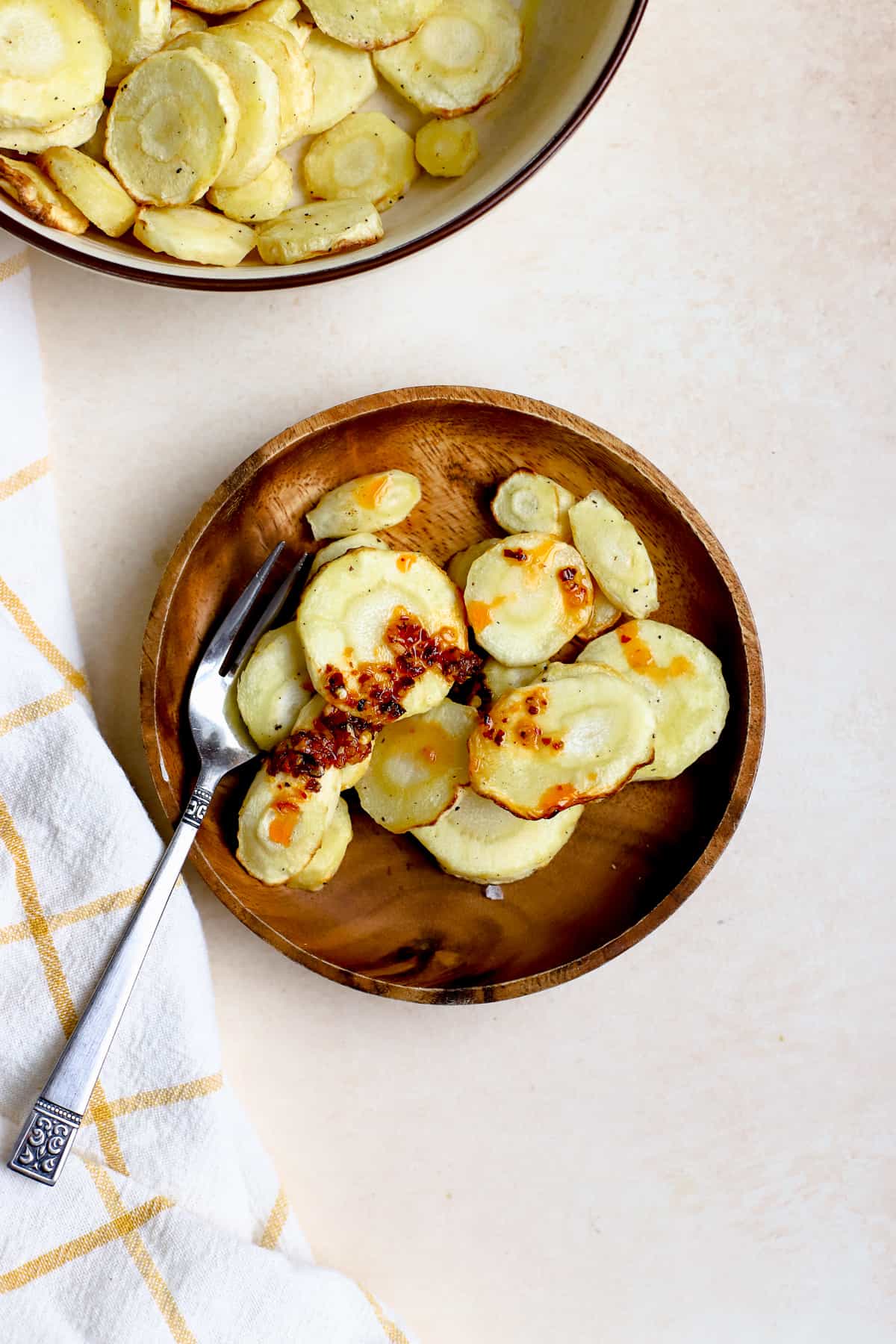 Air fryer parsnips on small wooden plate with fork topped with chili crunch and flaky salt, and other air fryer parsnips in beige serving bowl on the side.