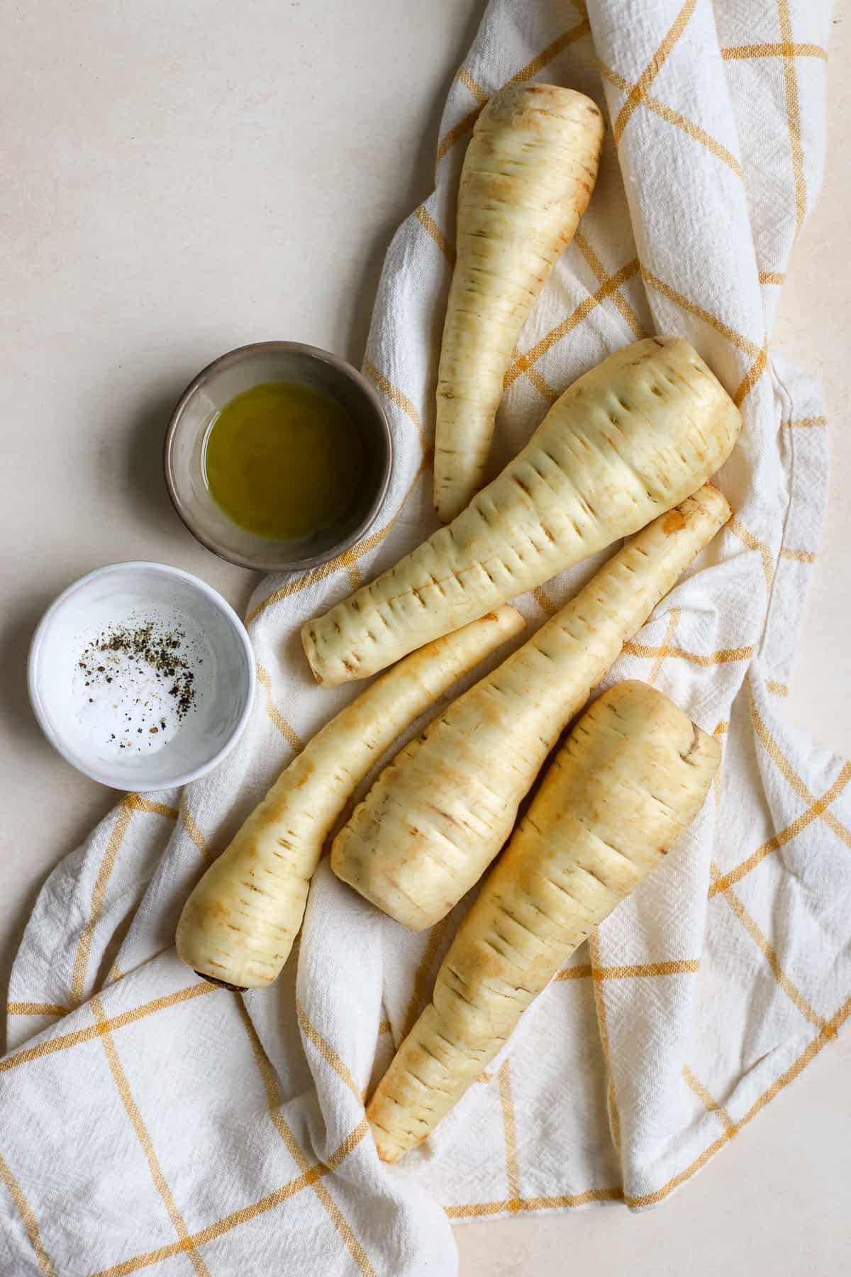 Whole raw parsnips on yellow and white checked linen with extra virgin olive oil, salt, and pepper on the side