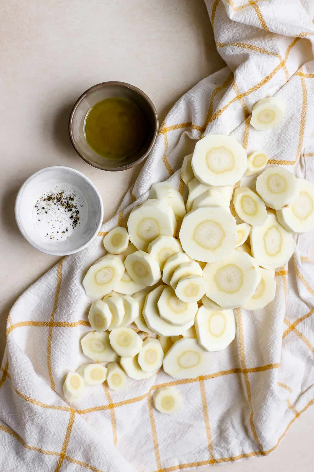 Peeled and sliced parsnips on yellow and white checked linen with extra virgin olive oil, salt, and pepper on the side.