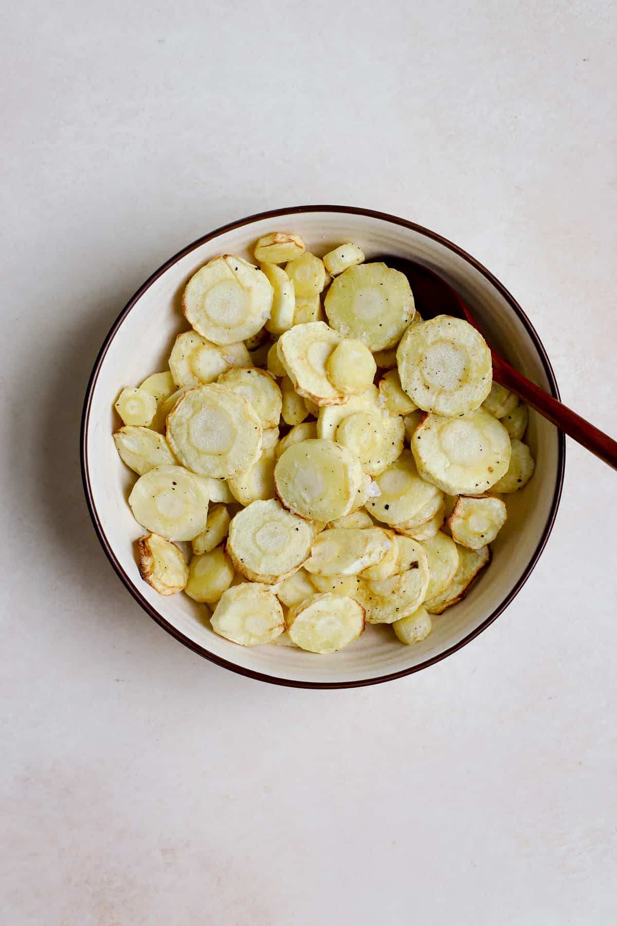 Air fryer parsnips in beige serving bowl with wooden spoon and yellow and white checked linen on the surface.