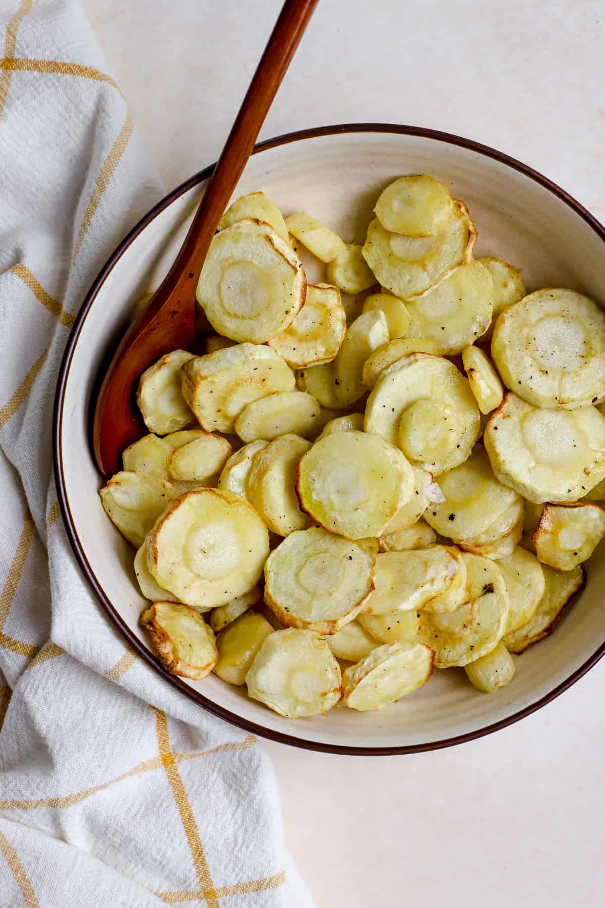Air fryer parsnips in beige serving bowl with wooden spoon and yellow and white checked linen on the surface.