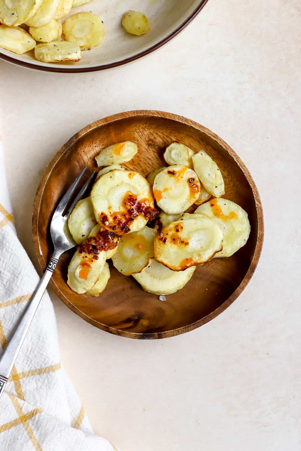 Air fryer parsnips on small wooden plate with fork topped with chili crunch and flaky salt, and other air fryer parsnips in beige serving bowl on the side.