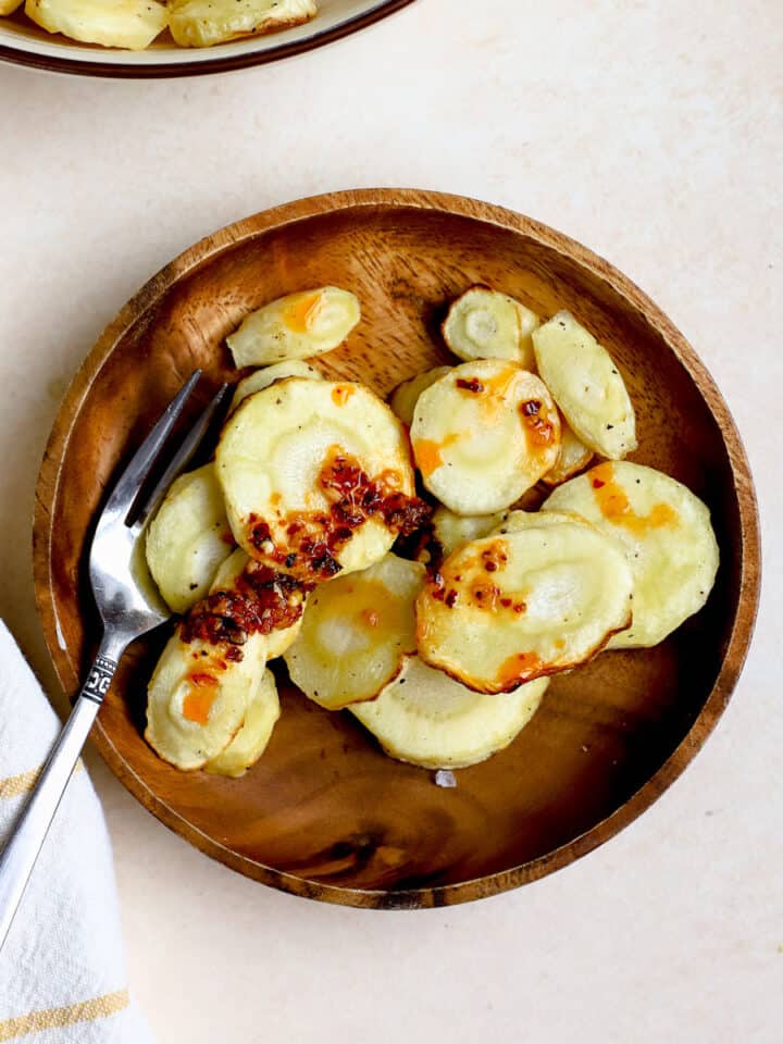 Air fryer parsnips on small wooden plate with fork topped with chili crunch and flaky salt, and other air fryer parsnips in beige serving bowl on the side