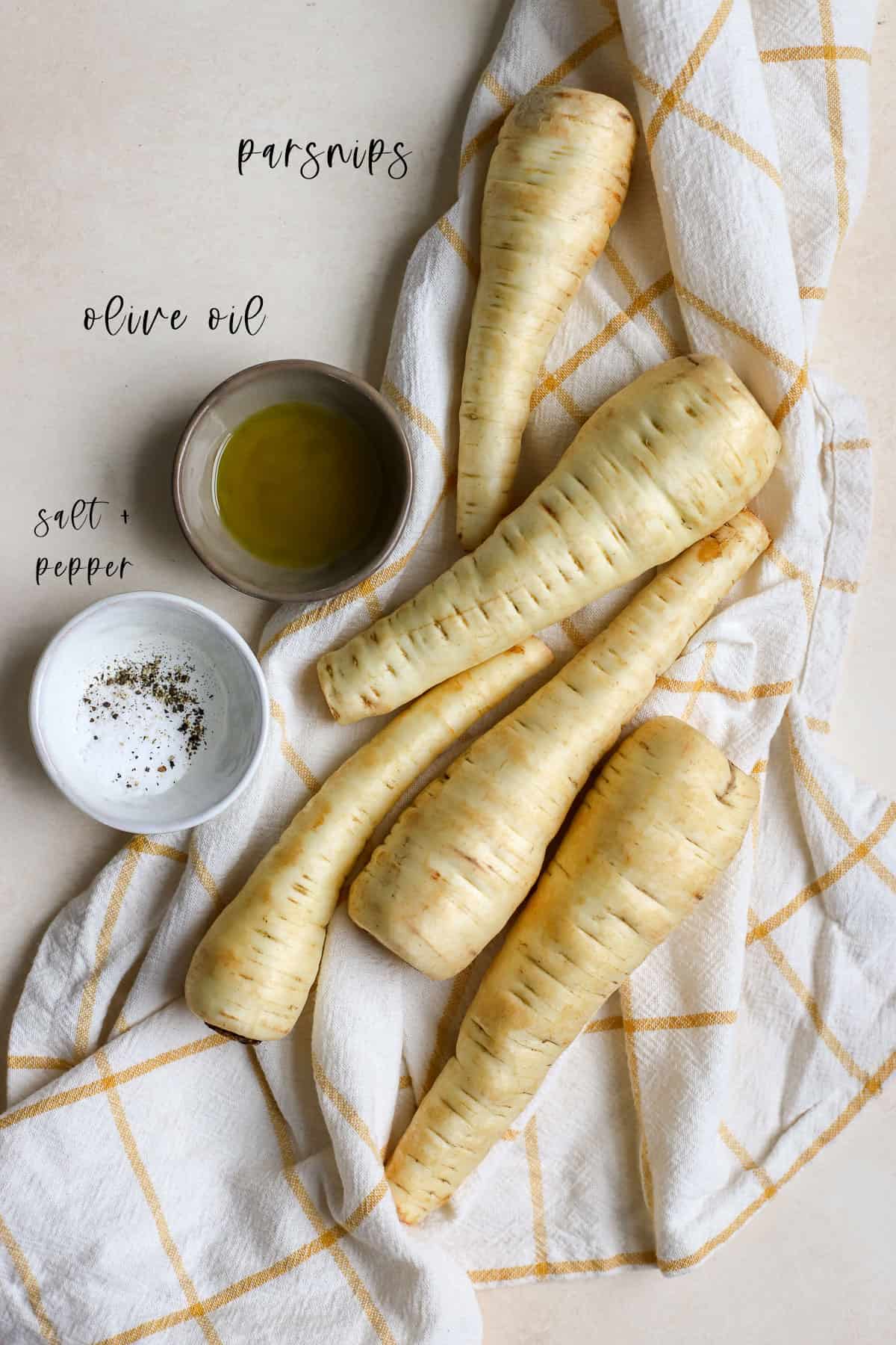 Whole raw parsnips on yellow and white checked linen with extra virgin olive oil, salt, and pepper on the side.