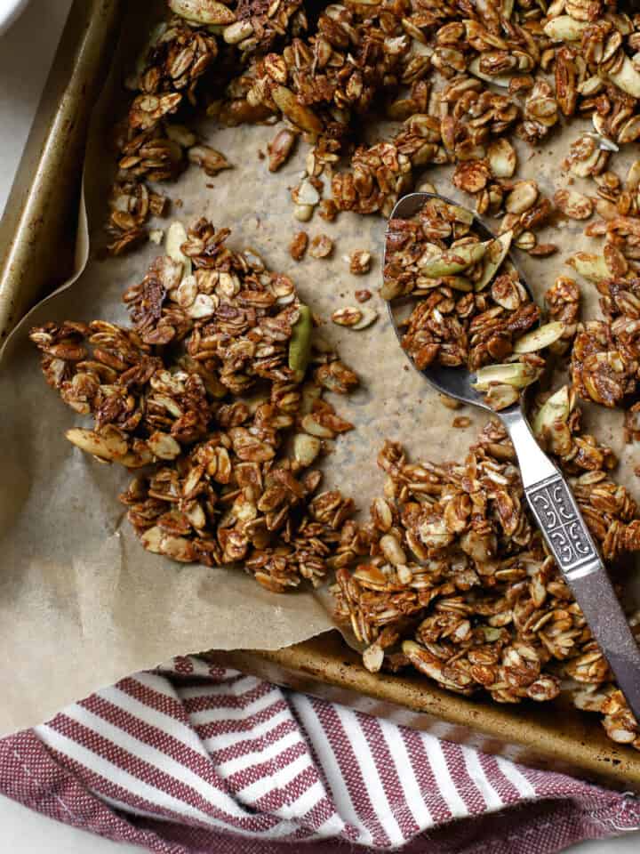 Ginger molasses granola crumbled on sheet pan lined with parchment paper, with spoon scooping some granola