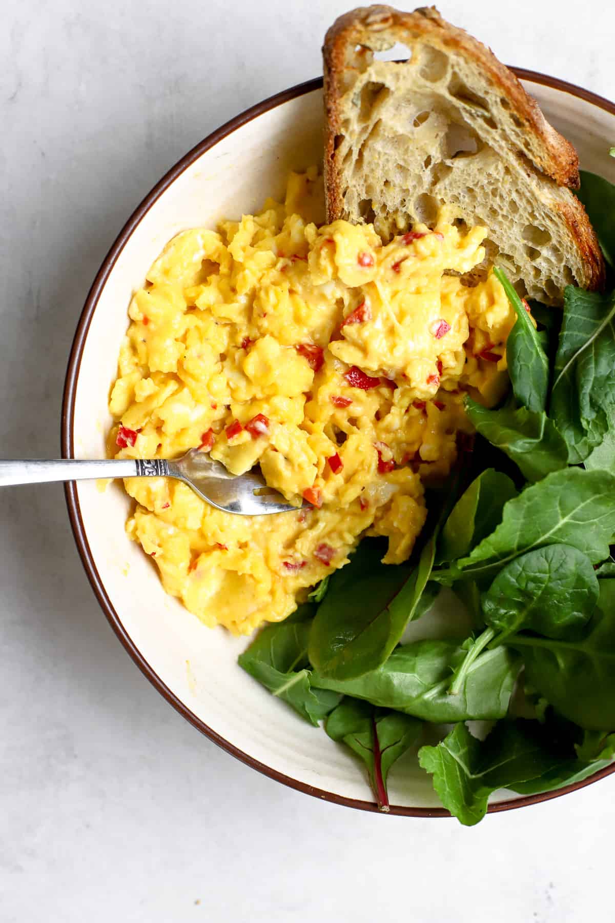 Large scoop of chilli scrambled eggs in beige bowl with pile of greens and toasted sourdough bread, with fork resting on side