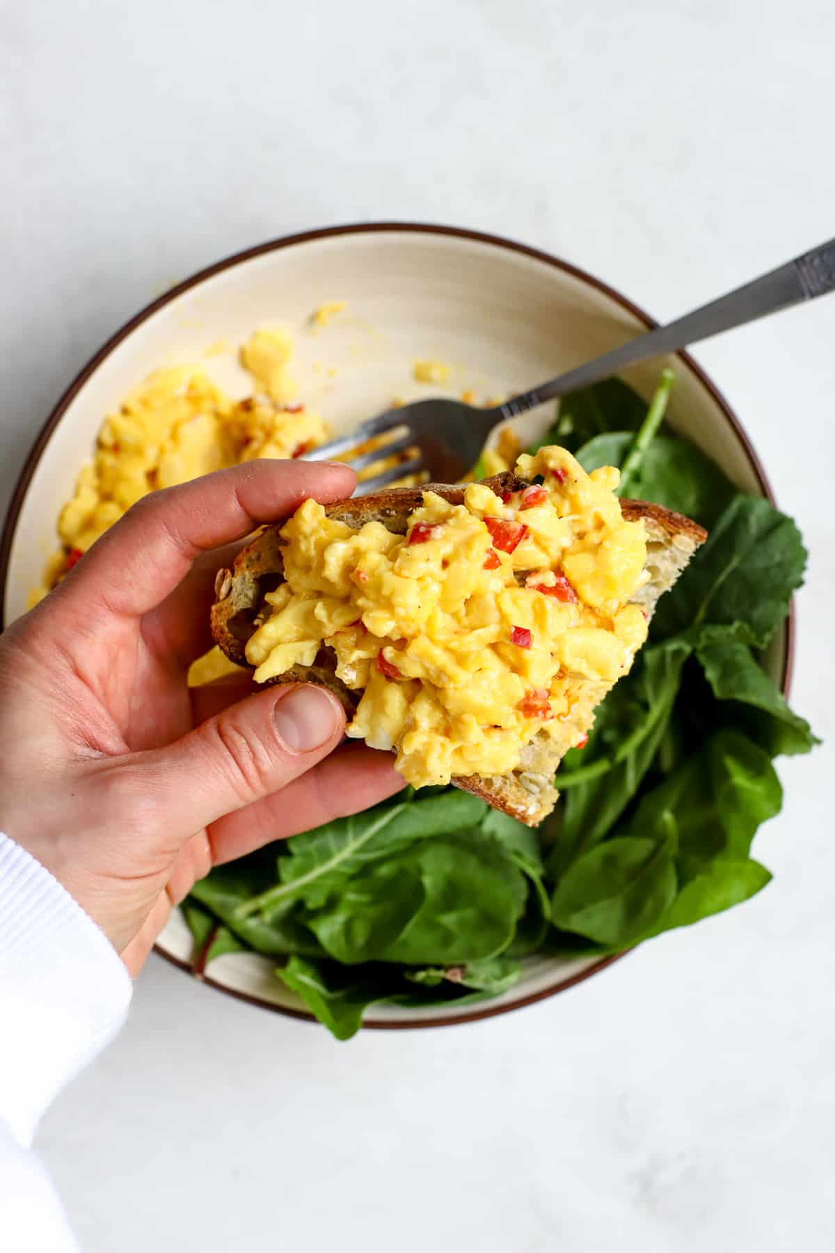 Chilli scrambled eggs scooped onto sourdough toast held by hand, with bowl of more eggs and greens in background.