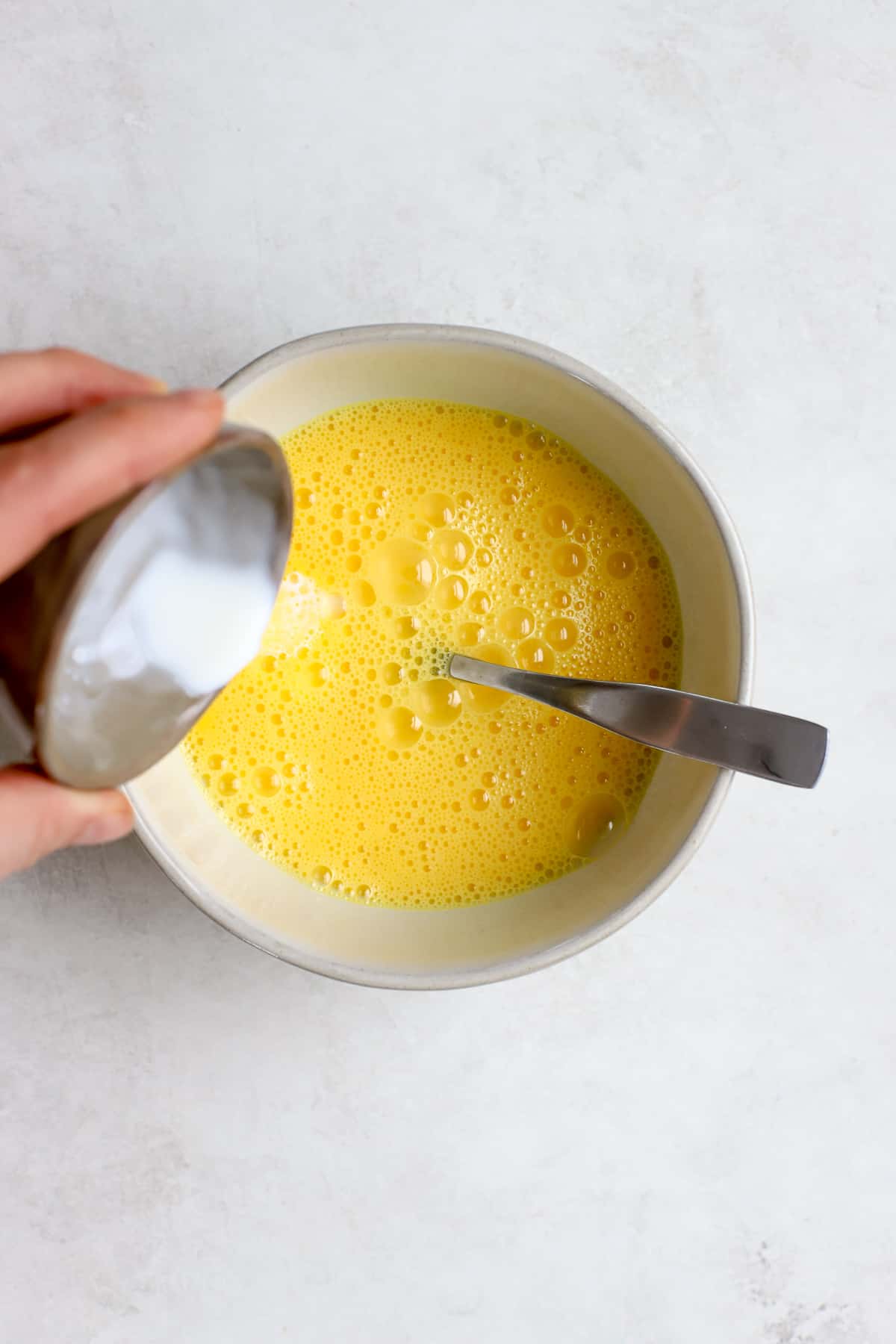 Whisked eggs in small white bowl with fork resting on side and cream being poured in.