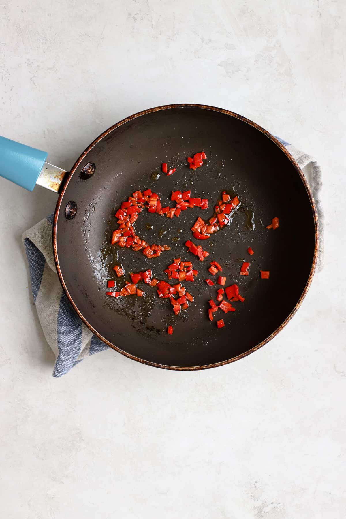 Finely diced red chilli in small blue sauté pan with olive oil, on white and blue striped towel, on light gray surface.