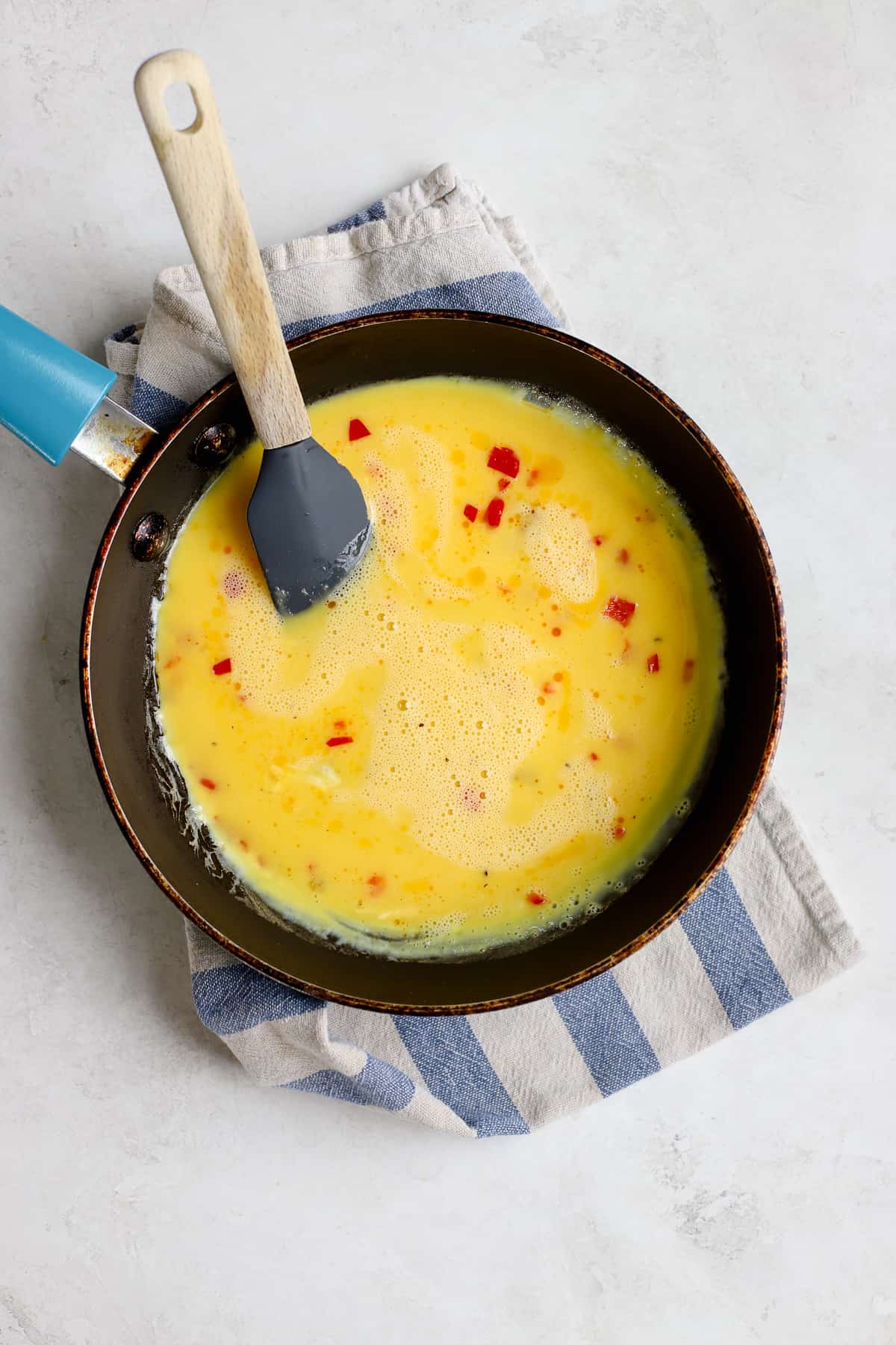 Egg mixture added to small blue sauté pan, just starting to cook, with small rubber spatula.