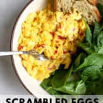 Large scoop of chilli scrambled eggs in beige bowl with pile of greens and toasted sourdough bread, with fork resting on side