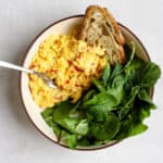 Large scoop of chilli scrambled eggs in beige bowl with pile of greens and toasted sourdough bread, with fork resting on side