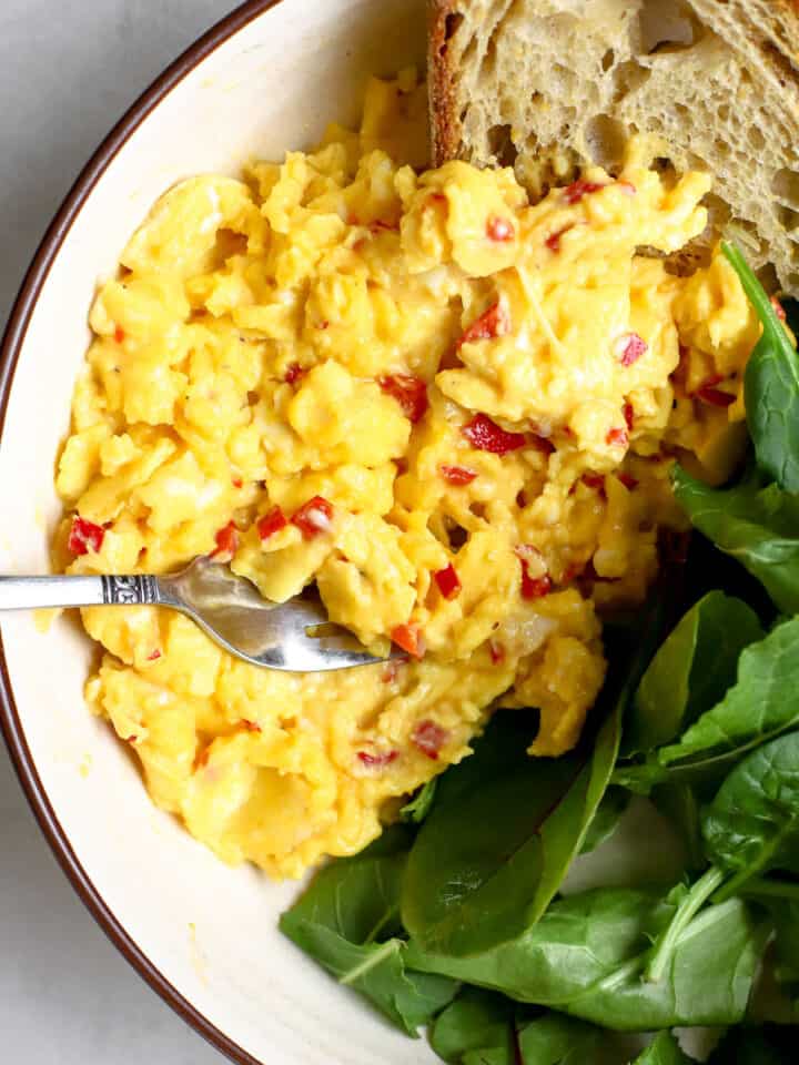 Large scoop of chilli scrambled eggs in beige bowl with pile of greens and toasted sourdough bread, with fork resting on side