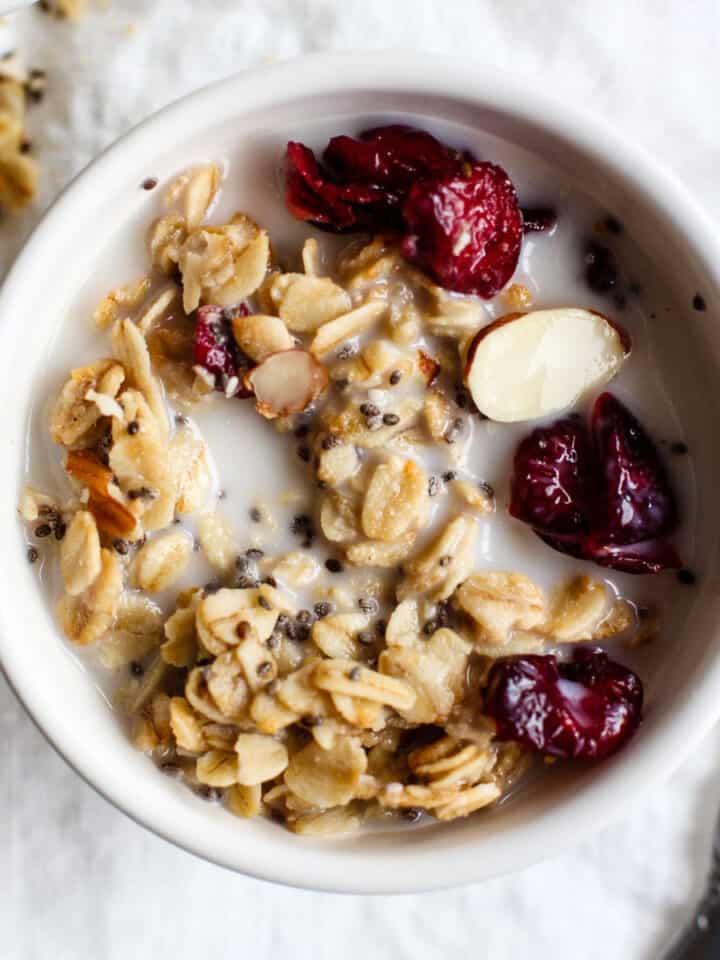 Maple cranberry almond granola in small white bowl with milk on white linen and silver spoon on side