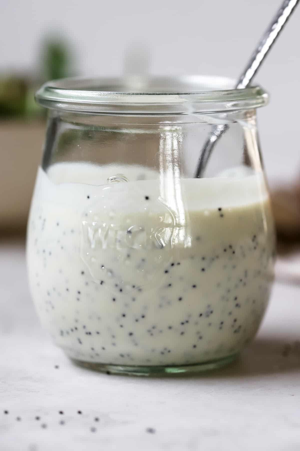 Tangy poppy seed dressing in small glass jar with spoon, on white and gray surface