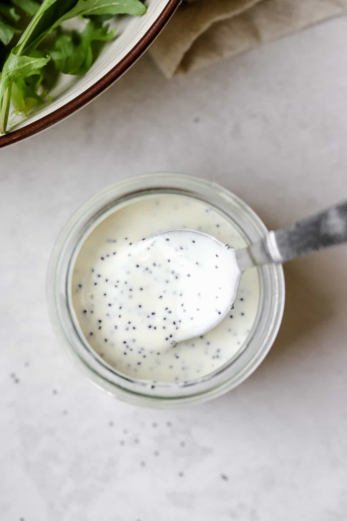 Poppy seed dressing in small glass jar with spoon scooping the dressing, and mixed greens salad in background on white and gray surface