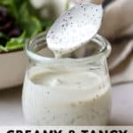 Tangy poppy seed dressing in small glass jar with spoon, on white and gray surface, with small wooden plate and bowl of mixed greens in the background