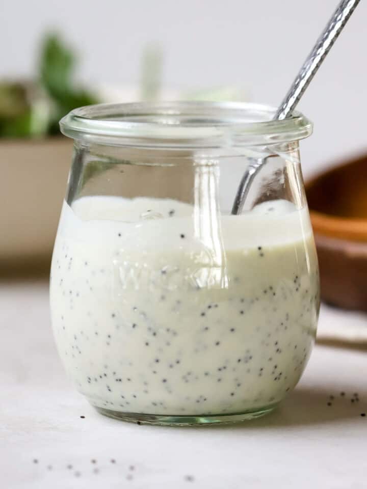 Tangy poppy seed dressing in small glass jar with spoon, on white and gray surface, with small wooden plate and bowl of mixed greens in the background