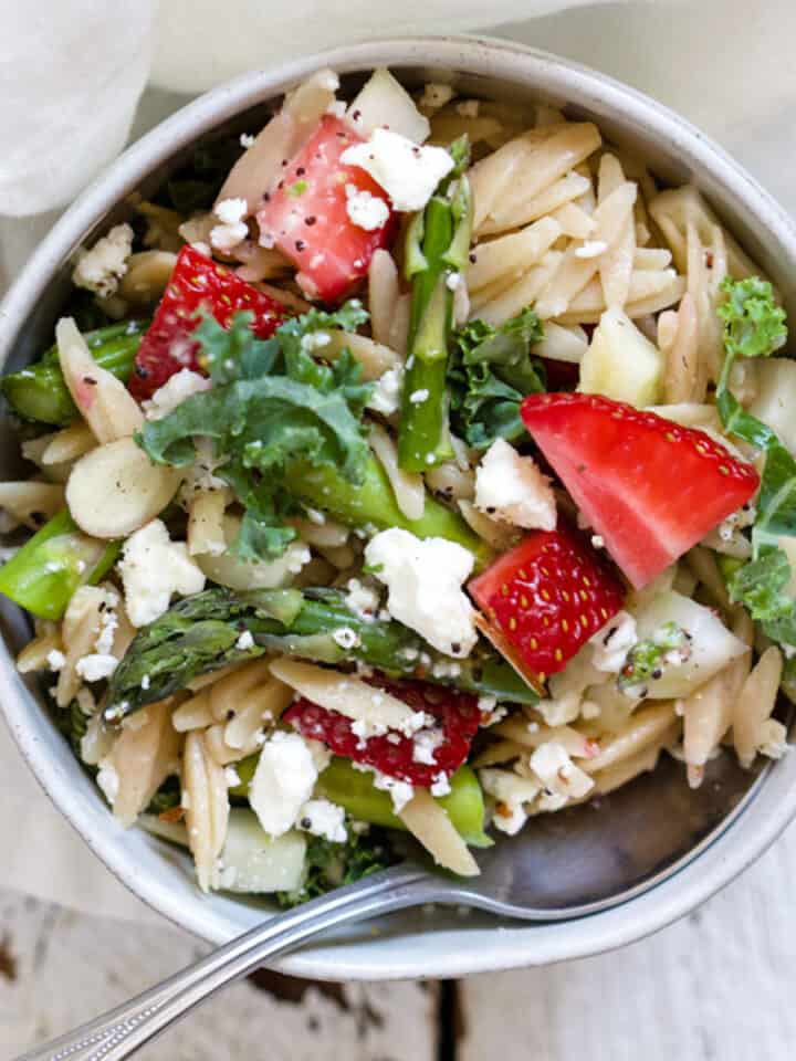 Strawberry feta orzo salad in white ceramic bowl with spoon, on white distressed wood surface