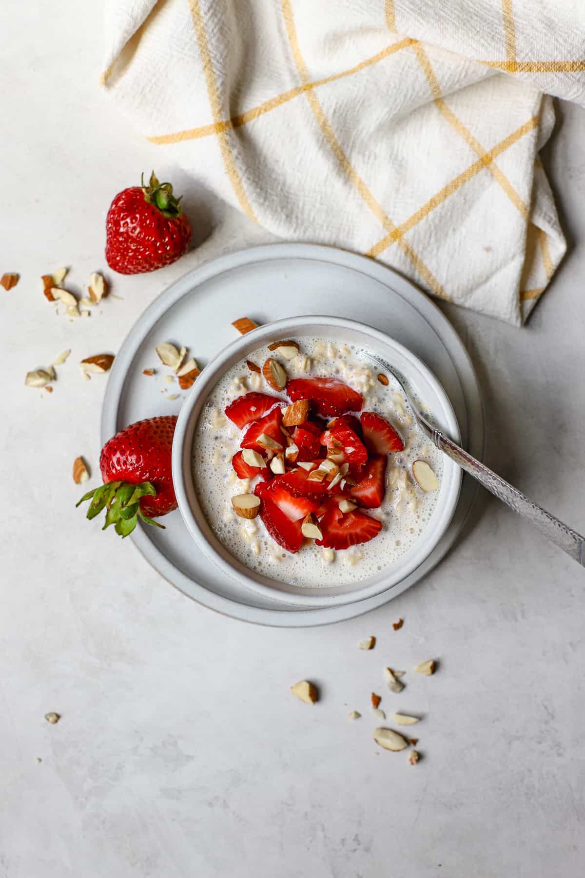 Strawberry shortcake overnight oats in light gray bowl on light gray plate, with chopped strawberries and almonds on top and on the side, on gray and white surface with white and yellow windowpane linen on the side