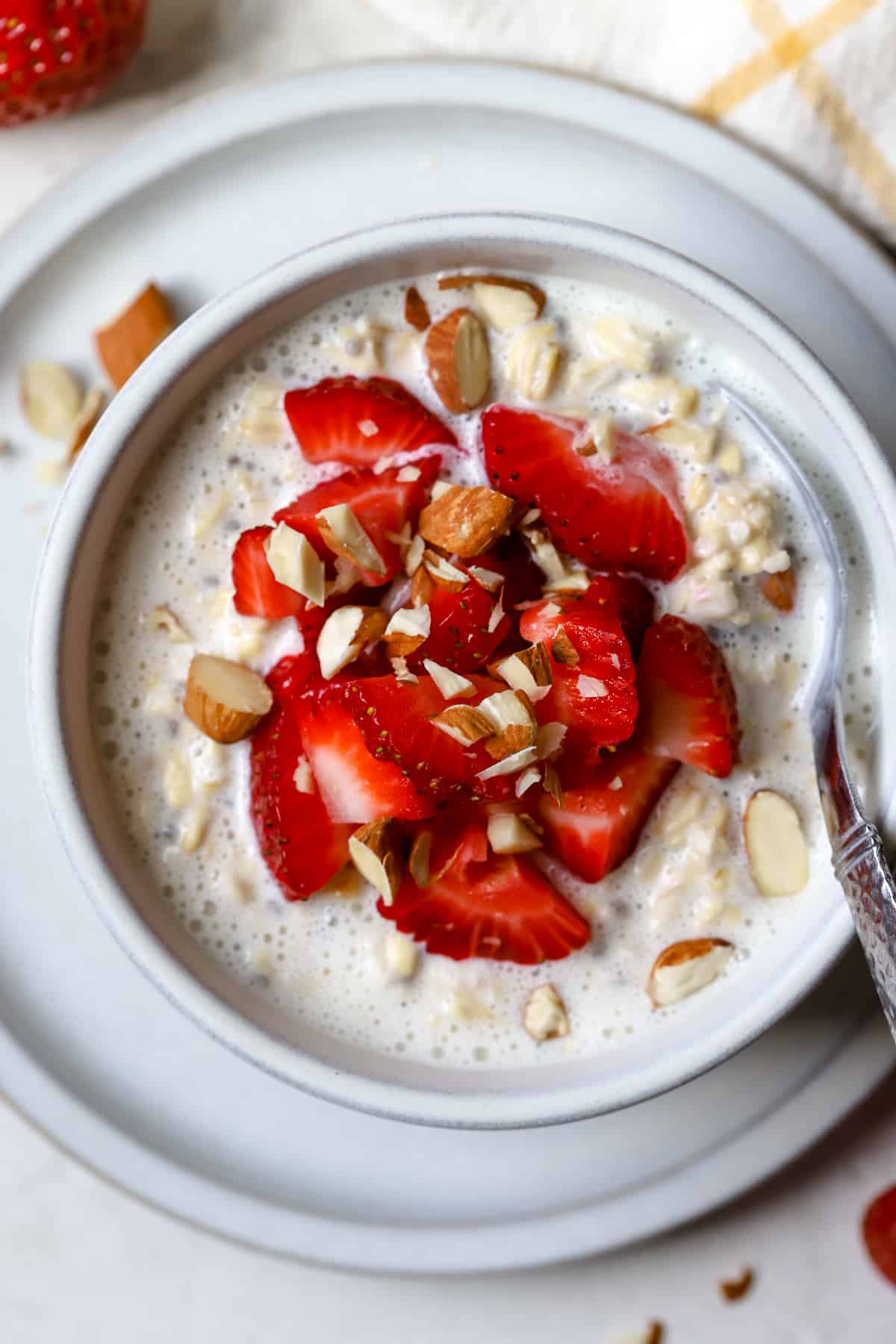 Strawberry shortcake overnight oats in light gray bowl on light gray plate, with chopped strawberries and almonds on top and on the side, on gray and white surface with white and yellow windowpane linen on the side