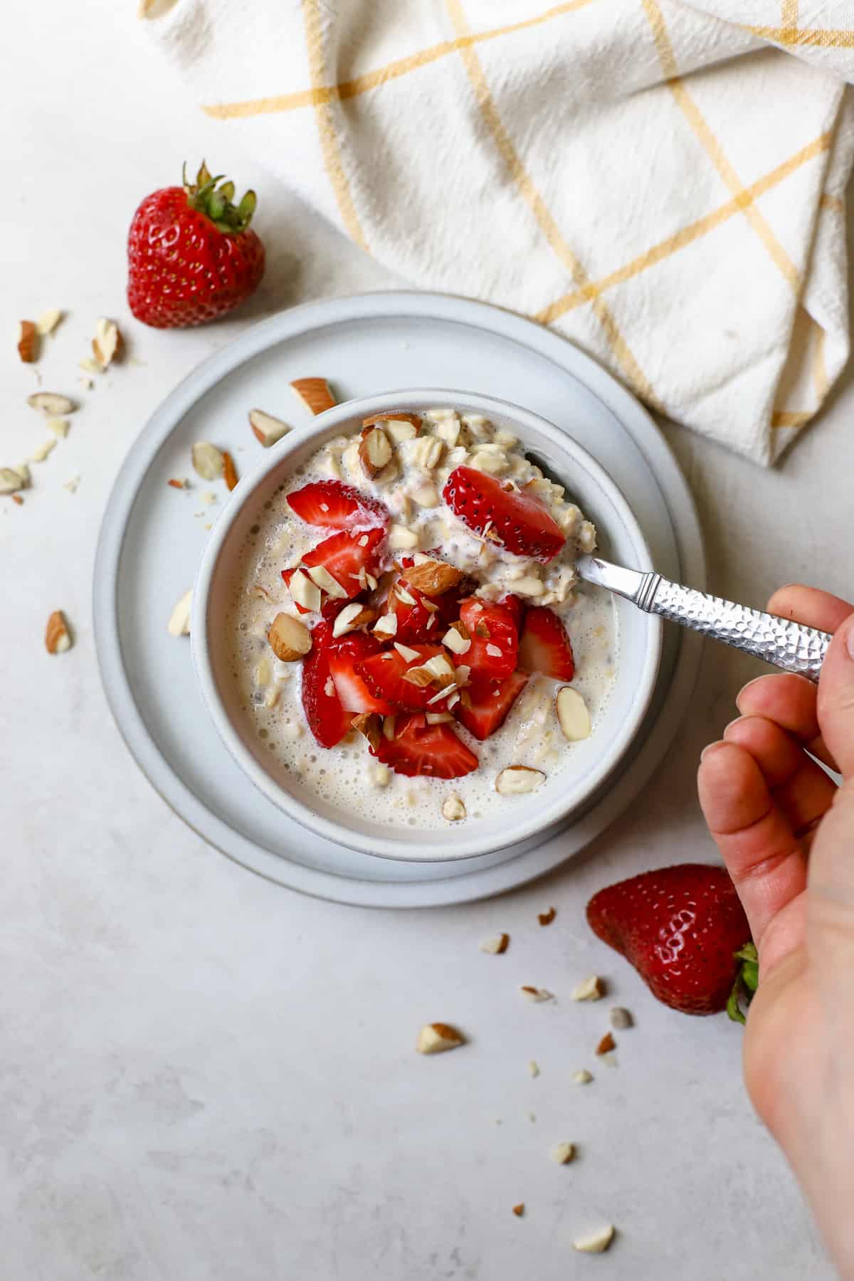 Strawberry shortcake overnight oats in light gray bowl on light gray plate, with chopped strawberries and almonds on top and on the side, hand holding silver spoon and scooping oats, on gray and white surface with white and yellow windowpane linen on the side