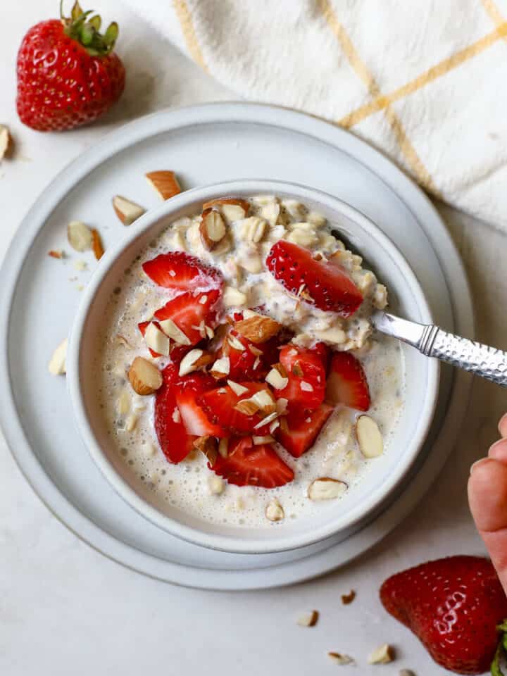 Strawberry shortcake overnight oats in light gray bowl on light gray plate, with chopped strawberries and almonds on top and on the side, hand holding silver spoon and scooping oats, on gray and white surface with white and yellow windowpane linen on the side