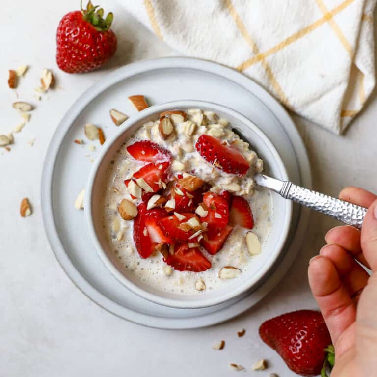 Strawberry shortcake overnight oats in light gray bowl on light gray plate, with chopped strawberries and almonds on top and on the side, hand holding silver spoon and scooping oats, on gray and white surface with white and yellow windowpane linen on the side