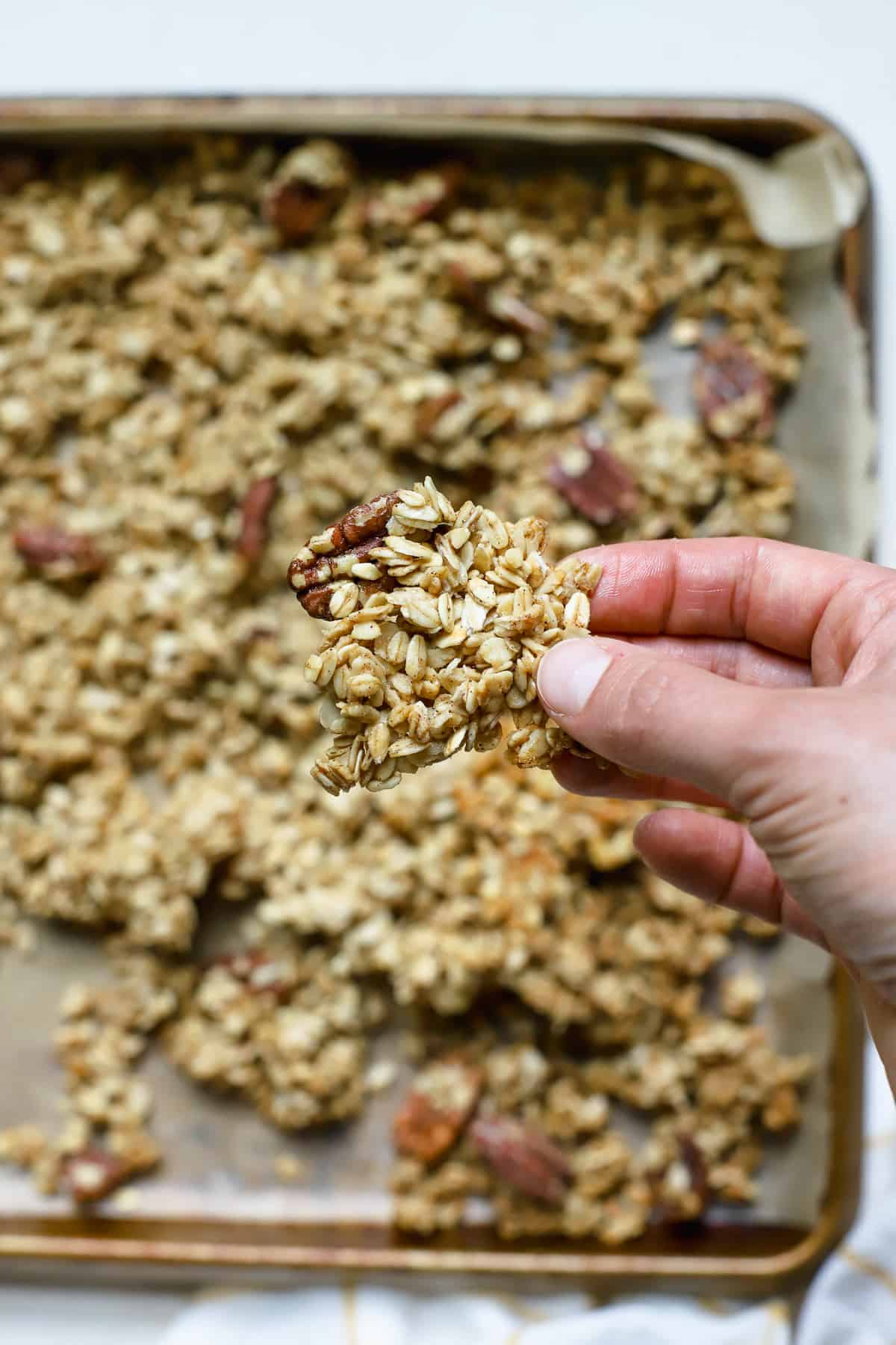 Fingertips holding a large cluster of maple pecan granola with sheet pan of granola in background.