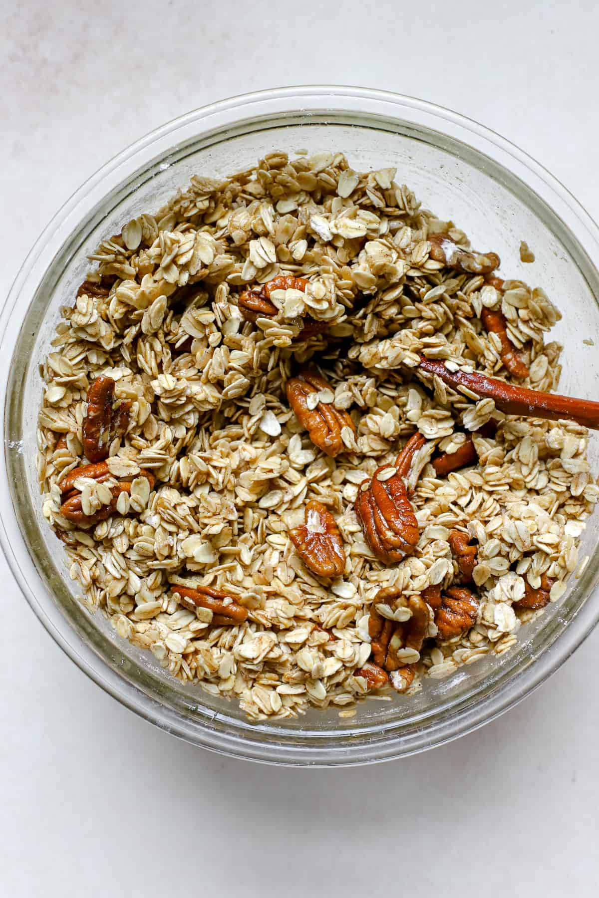 All maple pecan granola ingredients mixed in clear glass bowl with wooden spoon, on beige surface.